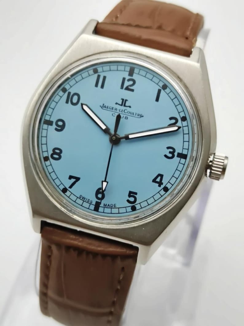 K&ouml;nnte beinhalten: Eine silberfarbene Armbanduhr mit hellblauem Zifferblatt und braunem Lederarmband. Das Zifferblatt zeigt schwarze Ziffern und den Markennamen "Jaeger-LeCoultre". Die Uhr hat ein rundes Zifferblatt und ein braunes Lederarmband.