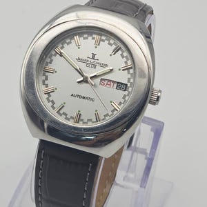 Puede incluir: Un reloj autom&aacute;tico Jaeger-LeCoultre Club plateado con esfera blanca, correa de cuero negra y la fecha mostrada como S&Aacute;B 28. El reloj tiene una esfera redonda y un bisel plateado. El reloj se muestra sobre un soporte transparente.