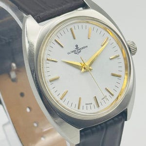 May include: A silver Ulysse Nardin wristwatch with a white face, gold hands, and a black leather strap. The watch face displays the brand name and an anchor logo. It is marked "Swiss Made".