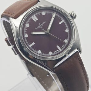 May include: A stainless steel Ulysse Nardin wristwatch with a burgundy dial and brown leather strap. The watch features white hour markers and hands, and a crown on the right side. The watch is marked "SWISS MADE".