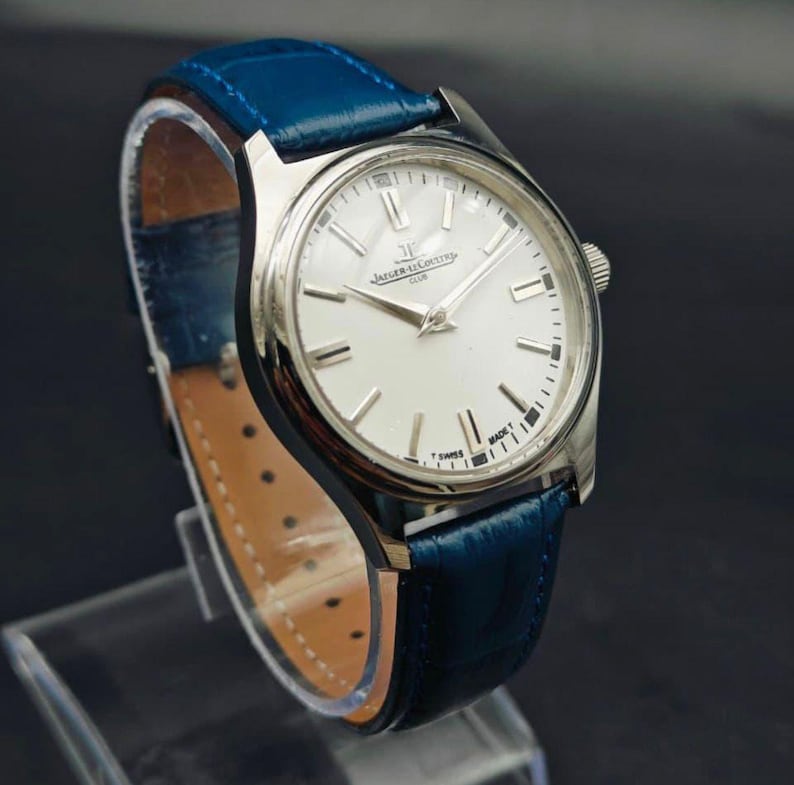 Peut inclure: Une montre-bracelet argent&eacute;e avec un cadran blanc et un bracelet en cuir bleu. Le cadran de la montre pr&eacute;sente des index et des aiguilles argent&eacute;s. Le bracelet est bleu vif, et la montre est expos&eacute;e sur un support transparent. La montre est marqu&eacute;e "Jaeger-LeCoultre Club" et "Swiss Made".