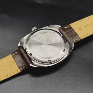May include: A silver-toned wristwatch with a circular case back featuring a brushed metal finish. The watch is attached to a dark brown leather strap with a light beige underside. The number 18 is printed on the strap near the watch casing.