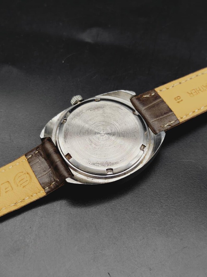 May include: A silver-toned wristwatch with a circular case back featuring a brushed metal finish. The watch is attached to a dark brown leather strap with a light beige underside. The number 18 is printed on the strap near the watch casing.