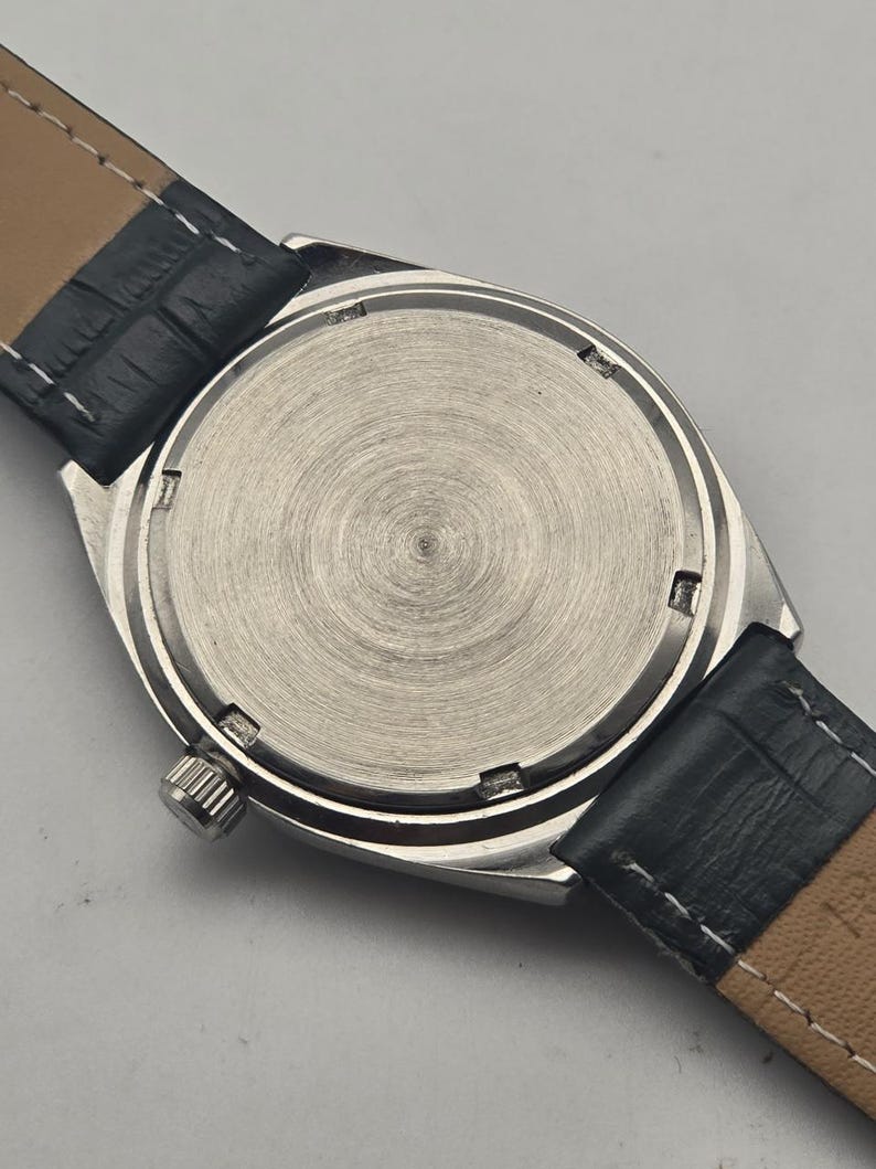 Puede incluir: Primer plano de la parte trasera de un reloj de pulsera plateado con una correa de cuero negro. La esfera del reloj es de color plata cepillada con un dise&ntilde;o circular. La correa de cuero negro est&aacute; cosida con hilo blanco.
