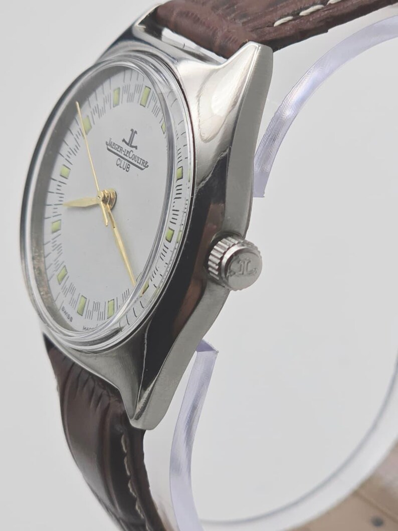 May include: A silver-toned Jaeger-LeCoultre Club watch with a white face, gold hands, and a brown leather strap. The watch face displays the Jaeger-LeCoultre logo and the word "CLUB". The watch is displayed on a clear stand.