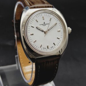 May include: A silver-tone Ulysse Nardin wristwatch with a white dial, silver hour markers, and silver hands. The watch features a brown leather strap with visible stitching and a crown on the right side. The dial displays the brand name and logo.