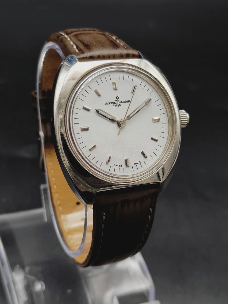 May include: A silver-tone Ulysse Nardin wristwatch with a white dial, silver hour markers, and silver hands. The watch features a brown leather strap with visible stitching and a crown on the right side. The dial displays the brand name and logo.