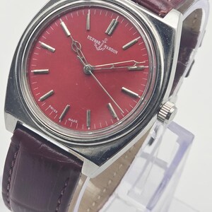 May include: A vintage Ulysse Nardin wristwatch with a red dial and silver-tone case. The watch features a burgundy leather strap. The dial has silver-tone hour markers and hands. The watch is marked "SWISS MADE".