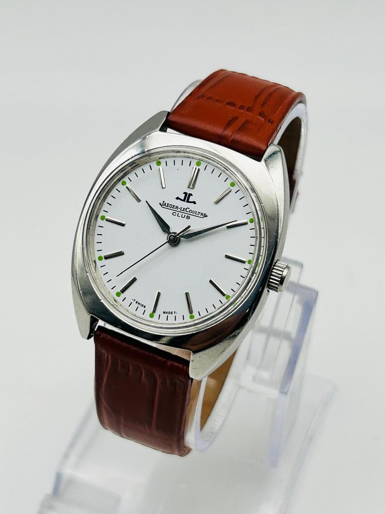 May include: A silver-toned Jaeger-LeCoultre Club watch with a white face, black hands, and a brown leather band. The watch face has green accents and the Jaeger-LeCoultre logo. The watch is displayed on a clear stand.