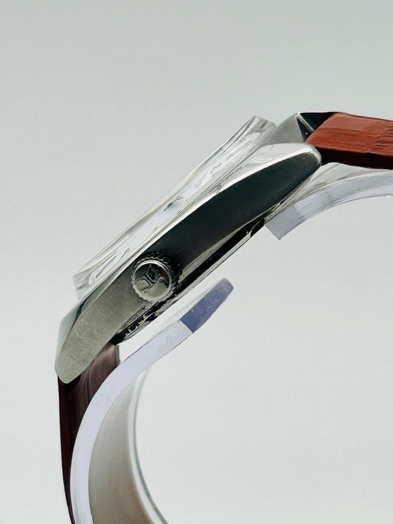 May include: A silver wristwatch with a brown leather band. The watch face is visible through a clear crystal. The watch has a round crown with a small emblem. The band is a dark brown color.