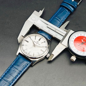 Peut inclure: Une montre-bracelet argent&eacute;e avec un cadran blanc et un bracelet en cuir bleu est mesur&eacute;e avec un pied &agrave; coulisse. La montre a des aiguilles et des marqueurs argent&eacute;s. Une jauge &agrave; cadran est &eacute;galement visible.