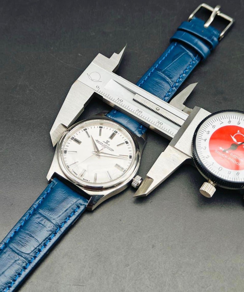 Peut inclure: Une montre-bracelet argent&eacute;e avec un cadran blanc et un bracelet en cuir bleu est mesur&eacute;e avec un pied &agrave; coulisse. La montre a des aiguilles et des marqueurs argent&eacute;s. Une jauge &agrave; cadran est &eacute;galement visible.
