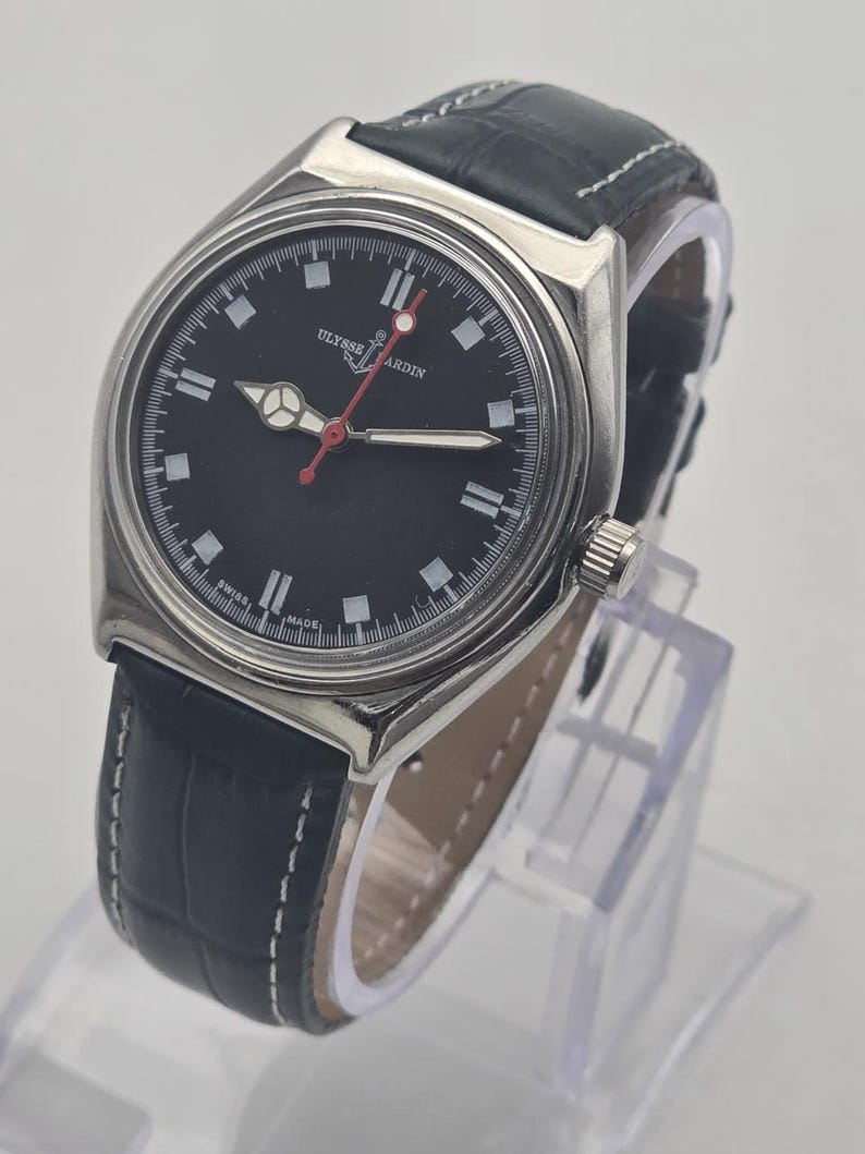 Puede incluir: Un reloj Ulysse Nardin de acero inoxidable con esfera negra y correa de cuero negro. El reloj tiene un bisel plateado, marcadores blancos y un segundero rojo. La correa tiene costuras blancas.