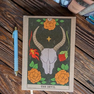 Könnte beinhalten: Ein Tarotkarten-Journal mit einem beigefarbenen Einband, der eine zentrale Illustration eines Schädels mit Hörnern, umgeben von orangefarbenen Rosen und dem Text "THE DEVIL" zeigt. Ein hellblauer Stift liegt neben dem Journal auf einer Holzoberfläche.