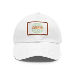 May include: White baseball cap with a curved brim. The cap features a rectangular patch with the word "CORNER" in a green and orange pattern, reminiscent of an eye chart, set against a white background, framed by a brown border.
