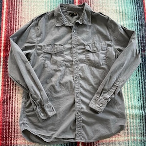 Puede incluir: Una camisa gris abotonada con dos bolsillos en el pecho y mangas largas. La camisa tiene un cuello clásico y puños abotonados. La tela parece ser una mezcla ligera de algodón o lino. La camisa está tendida sobre una superficie estampada.