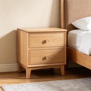 May include: A light oak wood bedside table featuring two stacked drawers with recessed paneling and small, round, metallic gold knobs. The piece is supported by four tapered legs and displays a natural wood grain finish throughout its structure.