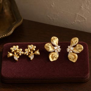 Puede incluir: Dos pares de pendientes dorados. El primer par presenta un grupo de pequeñas flores con piedras claras en el centro. El segundo par consiste en pendientes más grandes con forma de pétalos texturizados y filas de pequeñas piedras brillantes.