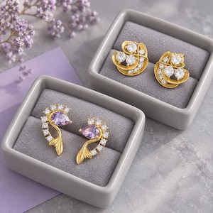 Puede incluir: Dos pares de pendientes dorados en cajas forradas de terciopelo gris. Un par presenta piedras redondas transparentes en diseño de espiral, mientras que el otro muestra piedras moradas en forma de pera con pequeños cristales curvos.