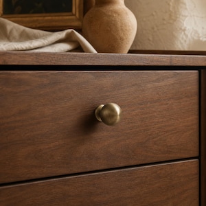 Op de afbeelding: Een ronde meubelknop van geborsteld messing is gecentreerd op een donkerbruin houten ladefront. De knop heeft een gladde, bolvormige vorm met een metallic afwerking, die contrasteert met de horizontale houtnerf van het paneel.