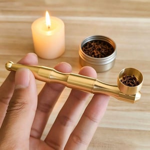 Puede incluir: Una pipa de metal de color dorado pulido con un tallo cilíndrico y un cuenco circular lleno de hierbas marrones secas. La pipa presenta un diseño liso y segmentado y se sostiene horizontalmente, mostrando un acabado metálico y sencillo.