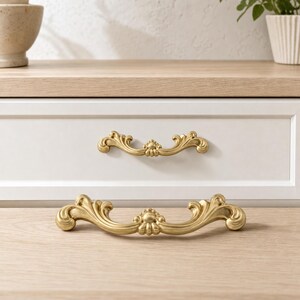 Pode incluir: Dois puxadores de gaveta em metal dourado com um design curvo e ornamentado, apresentando volutas simétricas e um elemento decorativo central. Um puxador está montado numa frente de gaveta branca, enquanto o outro repousa numa superfície de madeira clara.