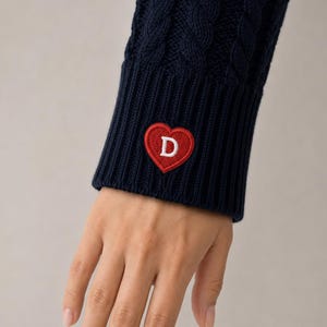 Può includere: Primo piano di una manica di maglione a trecce blu navy. Una toppa rossa a forma di cuore con una "D" bianca ricamata è cucita sul polsino. Il maglione è realizzato in un materiale spesso e strutturato.
