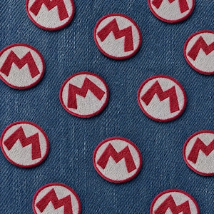 May include: Circular embroidered patches with a red "M" design within a white circle and a red border. The patches are arranged on a blue denim fabric background. These patches are suitable for customising clothing or accessories.