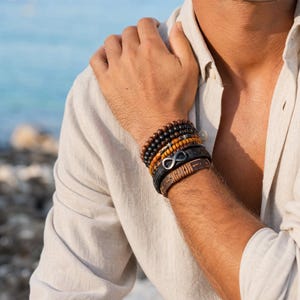 Könnte beinhalten: Ein Stapel aus fünf Armbändern am Handgelenk, bestehend aus dunkelbraunen und schwarzen runden Perlen, einem strukturierten beigen und schwarzen Kordelband, einem schwarzen geflochtenen Lederband mit silbernem Unendlichkeitssymbol und einer glatten beigen Ledermanschette.