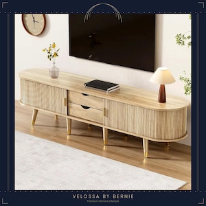 Può includere: Una console multimediale in legno chiaro dalla forma ovale arrotondata, dotata di due armadietti laterali con ante a listelli verticali, due cassetti centrali con piccole maniglie dorate e quattro gambe affusolate con piedini dorati.
