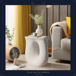 Peut inclure: Une table d'appoint blanche et sculpturale, avec un design arrondi unique et une grande ouverture centrale. La table est tachetée et supporte un vase blanc avec de la verdure, un livre et une tasse. Collection "Velossa by Bernie".