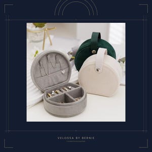 May include: A gray, round jewelry box with an open lid displaying necklaces and rings. Two other round jewelry cases, one cream-colored and one dark green, are also visible. The cases have handles. The text "VELOSSA BY BERNIE" is at the bottom.