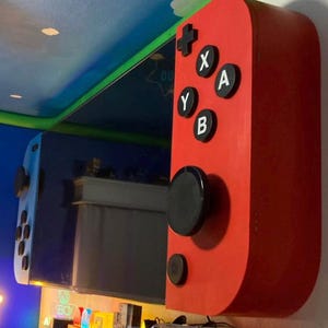 May include: A large, red, wall-mounted controller with black buttons and a black joystick. The controller has the letters X, A, Y, and B on it. A blue and white controller is also visible.