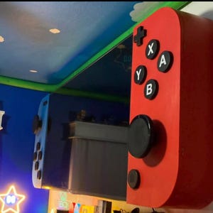 May include: A large, red, rectangular wall-mounted controller accessory featuring a black circular joystick, a small black circular button, and four black buttons labeled with the white letters X, A, Y, and B, plus a black directional pad.