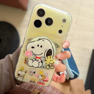 May include: A clear yellow phone case featuring Snoopy and Woodstock characters. The case has a pop-up grip with Snoopy holding a flower and Woodstock. Additional Snoopy and Woodstock graphics are visible on the case. The case is designed for a smartphone.