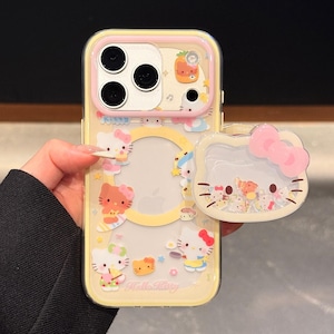 Peut inclure: Une coque de t&eacute;l&eacute;phone jaune avec une bordure rose et un pop-socket assorti. La coque pr&eacute;sente des personnages Hello Kitty et d'autres illustrations mignonnes. Le pop-socket a un design de n&oelig;ud rose. Le texte "Hello Kitty" est visible.