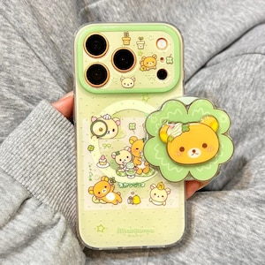 Peut inclure: Une coque de téléphone vert clair avec un motif d'ours de dessin animé et un pop socket assorti. La coque présente des illustrations d'ours, de plantes et du texte. Le pop socket a un visage d'ours avec un motif de fleur.