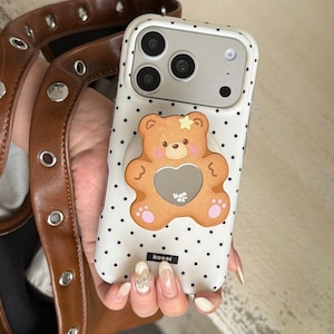 May include: White phone case with black polka dots and a brown teddy bear design. The bear has a heart-shaped cutout and a star on its head. The case has a camera cutout and the word "koool" is printed on the bottom.