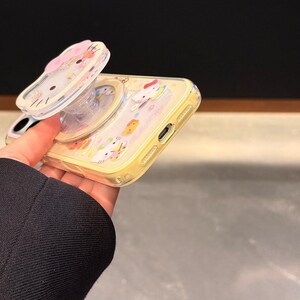 Peut inclure: Coque de t&eacute;l&eacute;phone jaune avec un support pop-up repr&eacute;sentant un personnage de dessin anim&eacute;. La coque a un dos transparent et une bordure jaune. Le support pop-up est transparent avec un motif de n&oelig;ud rose. Le texte "Super Cute Case" est en bas &agrave; droite.