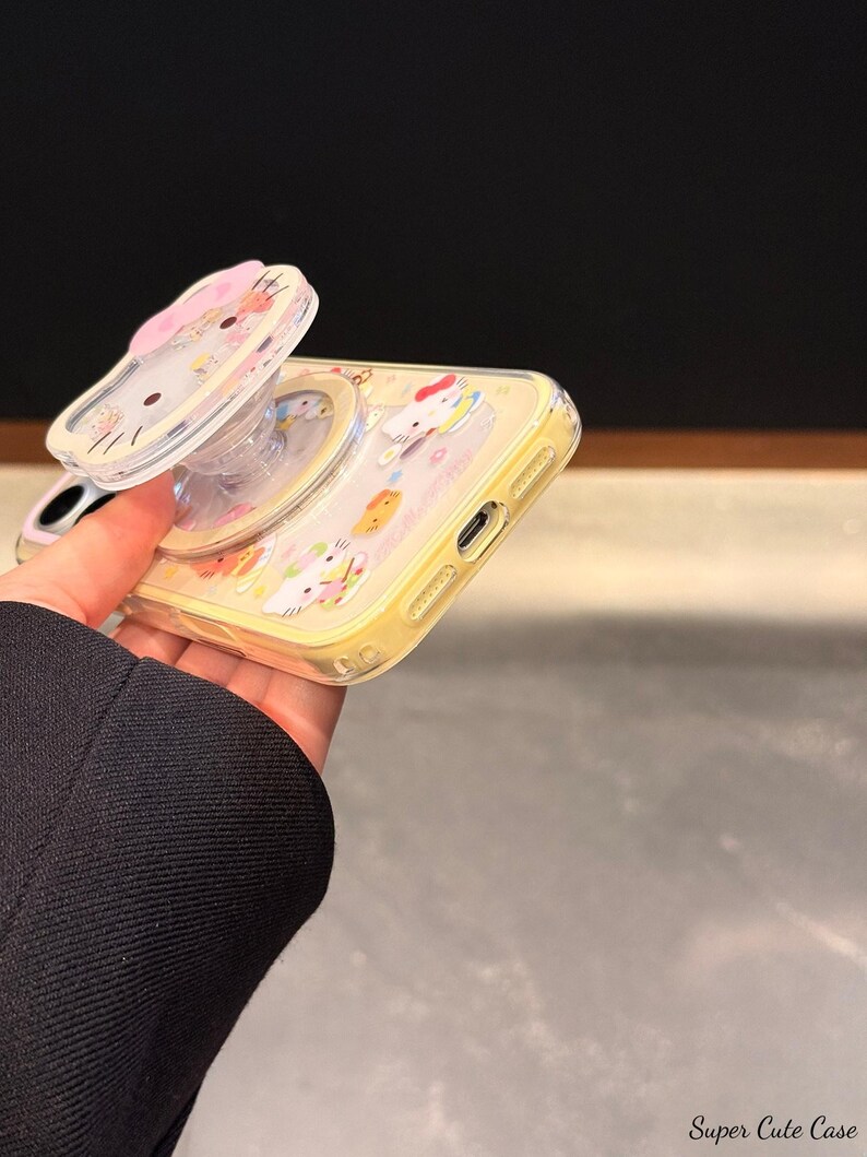 Peut inclure: Coque de t&eacute;l&eacute;phone jaune avec un support pop-up repr&eacute;sentant un personnage de dessin anim&eacute;. La coque a un dos transparent et une bordure jaune. Le support pop-up est transparent avec un motif de n&oelig;ud rose. Le texte "Super Cute Case" est en bas &agrave; droite.