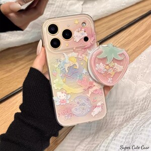 May include: A clear phone case with a colorful, whimsical design featuring cartoon characters, stars, and strawberries. A pink strawberry-shaped phone grip is attached. The text "Super Cute Case" is visible.