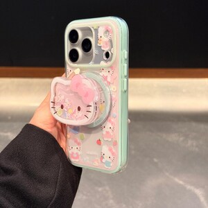 May include: Clear phone case with a pink and mint green border, featuring a Hello Kitty design. The case has a pop-up grip with a Hello Kitty face and bow. The case is decorated with Hello Kitty characters and small fruit illustrations.