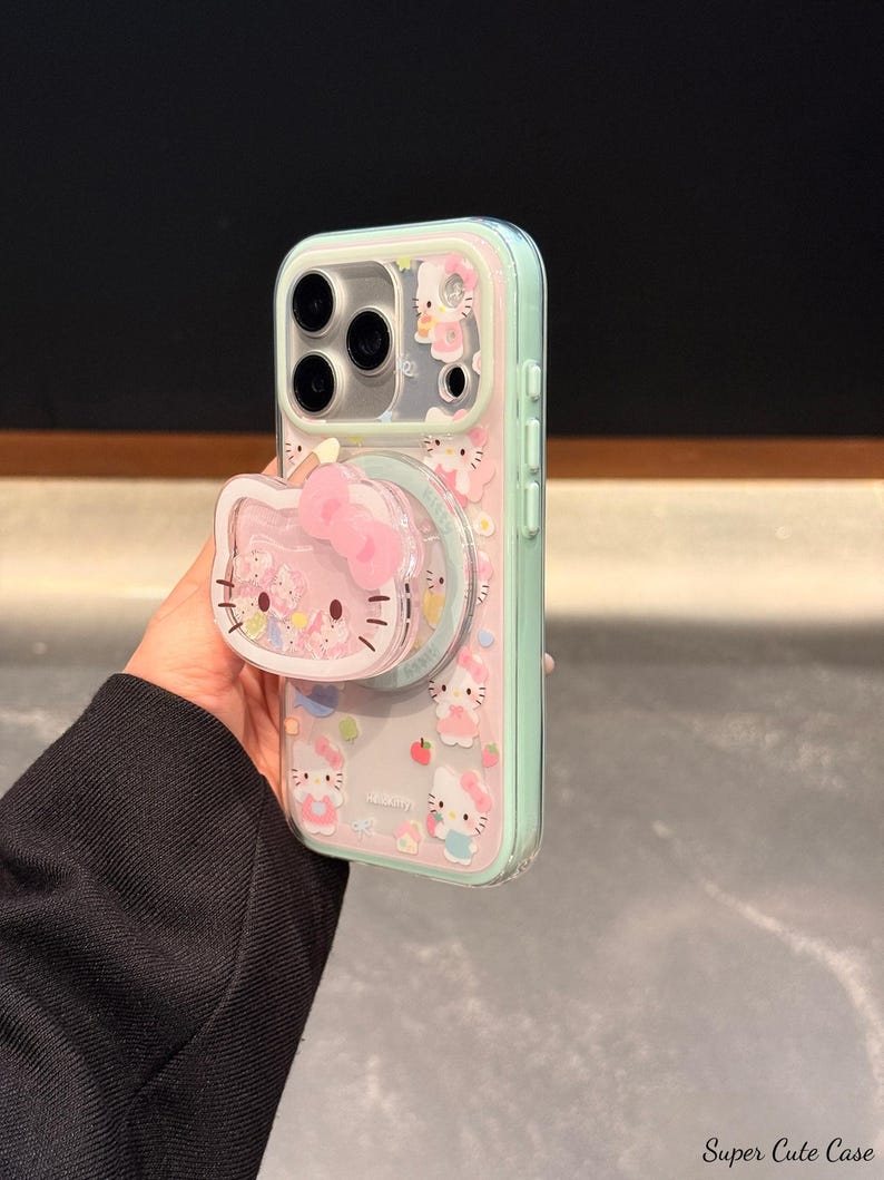 May include: Clear phone case with a pink and mint green border, featuring a Hello Kitty design. The case has a pop-up grip with a Hello Kitty face and bow. The case is decorated with Hello Kitty characters and small fruit illustrations.