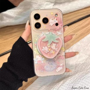 May include: A clear phone case with a pink strawberry design and various cartoon characters. The case has a pop-up strawberry grip and is decorated with stars and other cute elements. The text "Super Cute Case" is visible at the bottom.