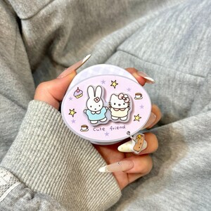 May include: A light purple phone grip featuring cartoon characters, a bunny and Hello Kitty, with the words "Cute friend". The grip has a small charm and is held against a gray background.