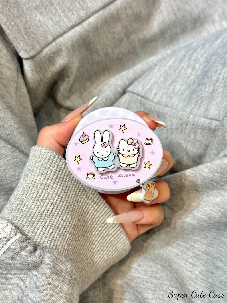 May include: A light purple phone grip featuring cartoon characters, a bunny and Hello Kitty, with the words "Cute friend". The grip has a small charm and is held against a gray background.