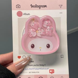 May include: A pink phone grip shaped like a cartoon character, with small, clear, decorative figures inside. The grip is on a card designed to look like an Instagram post, with the word "Instagram" at the top.