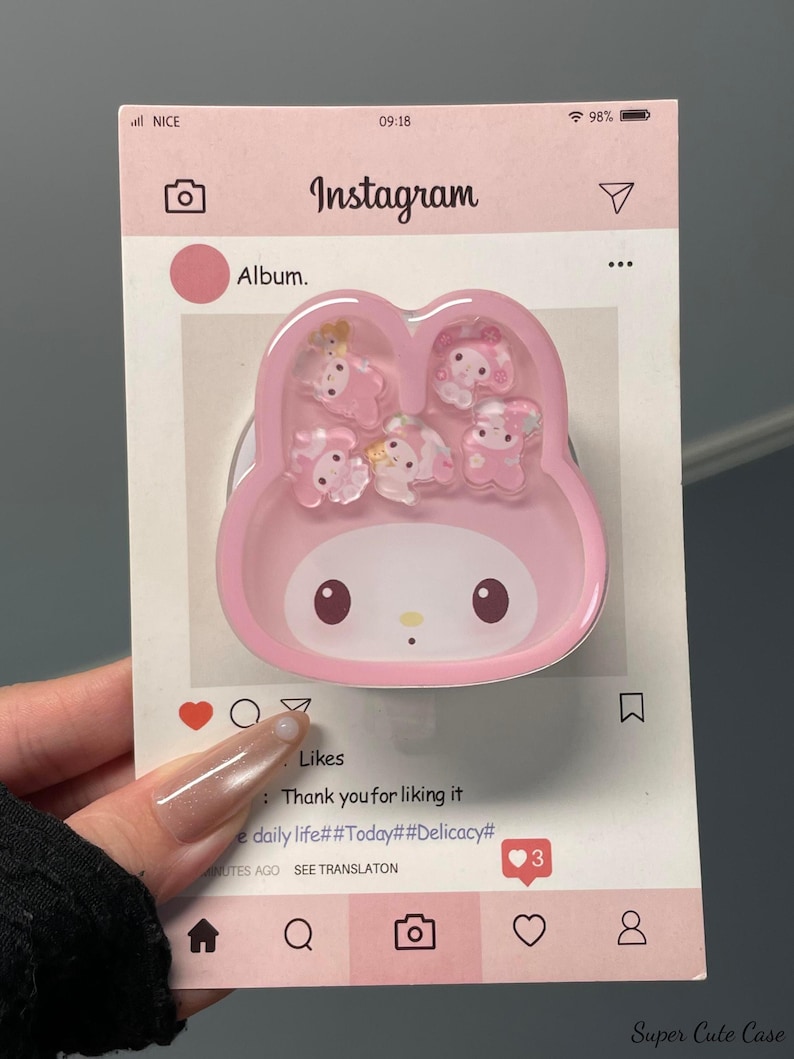 May include: A pink phone grip shaped like a cartoon character, with small, clear, decorative figures inside. The grip is on a card designed to look like an Instagram post, with the word "Instagram" at the top.