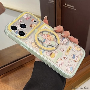 以下が含まれることがあります： 薄い緑色の縁と黄色のアクセントが付いた透明な携帯電話ケース。ケースには、さまざまなキャラクターの漫画デザインと、星型のデザインの黄色のリングホルダーが付いています。「chikawa」という文字がケースに表示されています。