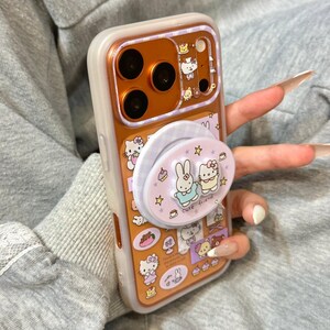 May include: A clear phone case with a gold-colored interior and a pop-up grip featuring cartoon characters. The case has a white border and is decorated with various cartoon images, including Hello Kitty and a bunny. The text "Super Cute Case" is visible.